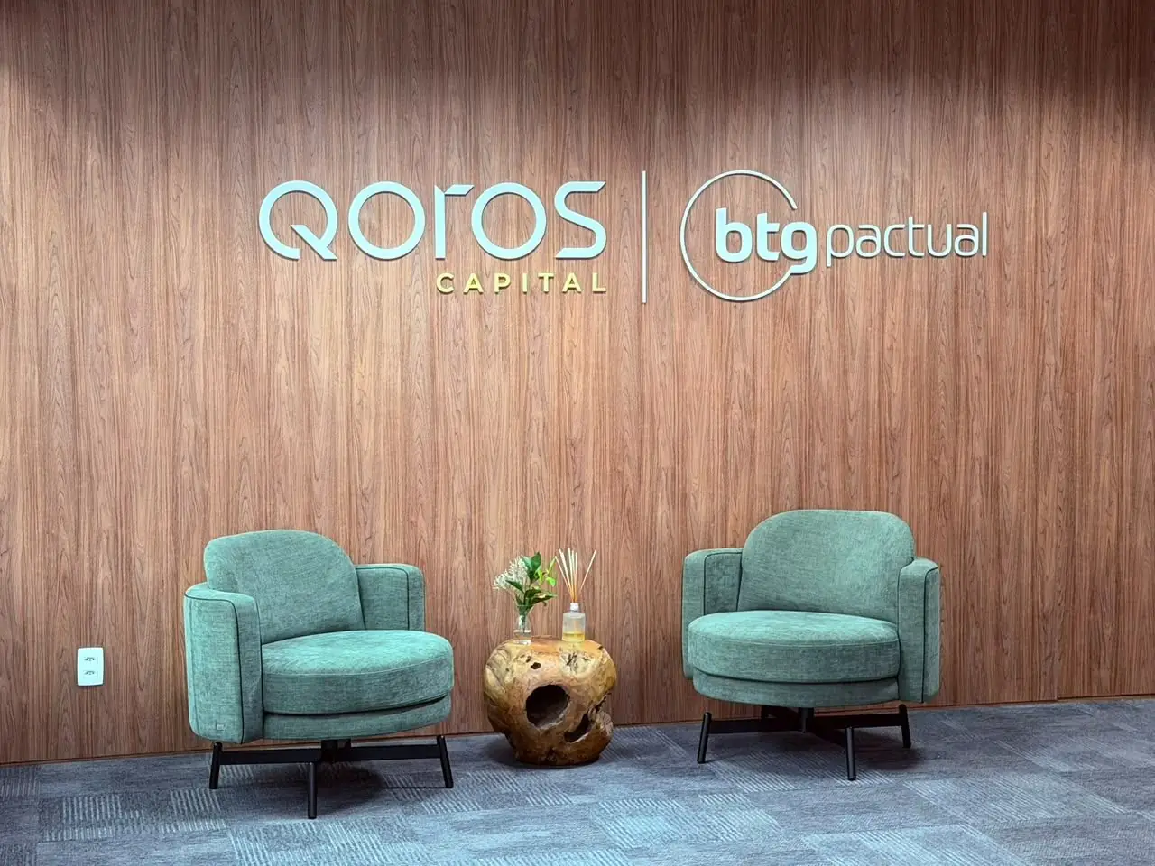 Recepção da Qoros Capital: painel de madeira com os logos Qoros Capital e BTG Pactual e duas poltronas